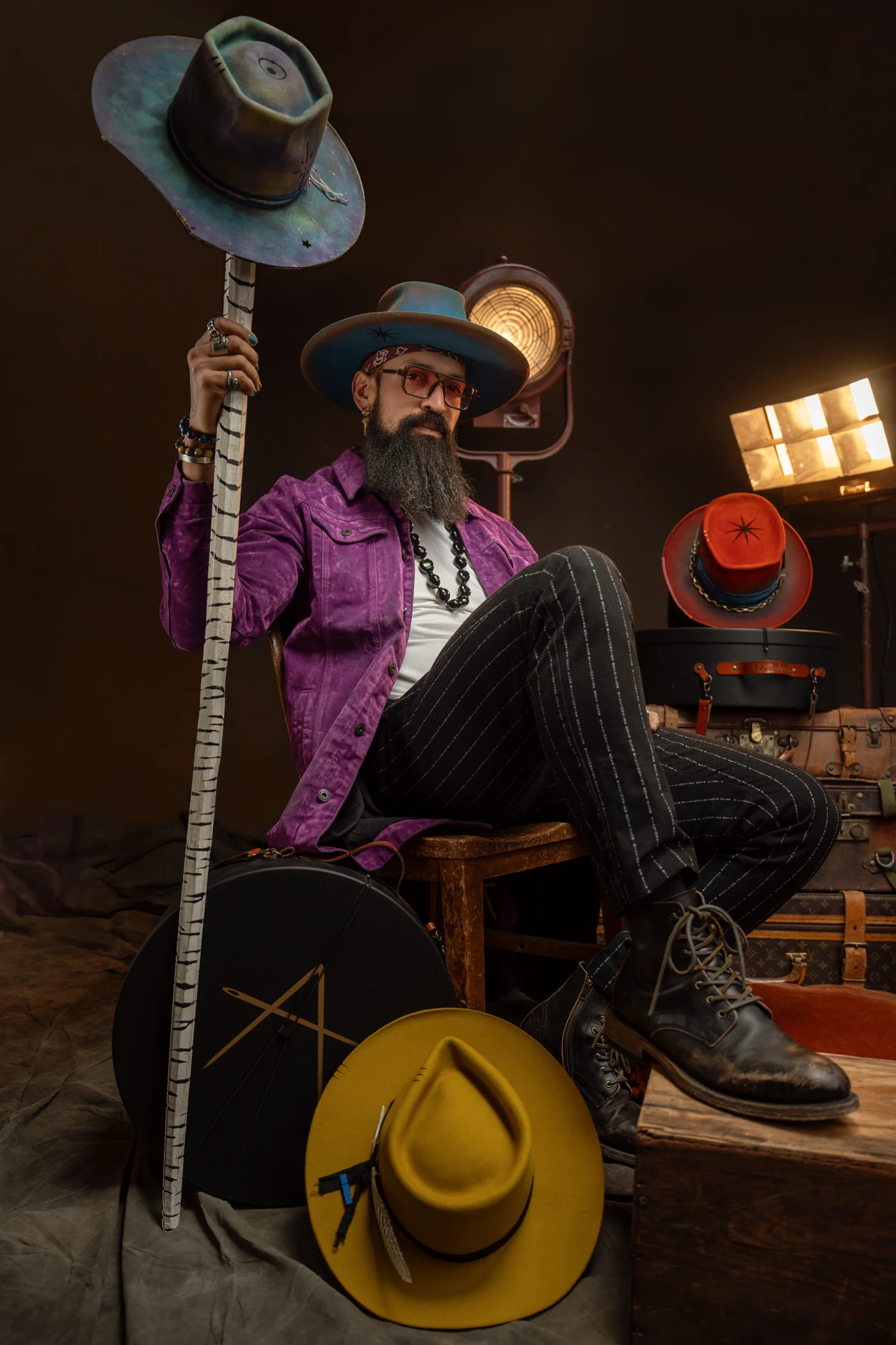 Full-length portrait of La Sombra in his atelier, wearing a purple jacket and boots, holding a sculptural hat aloft